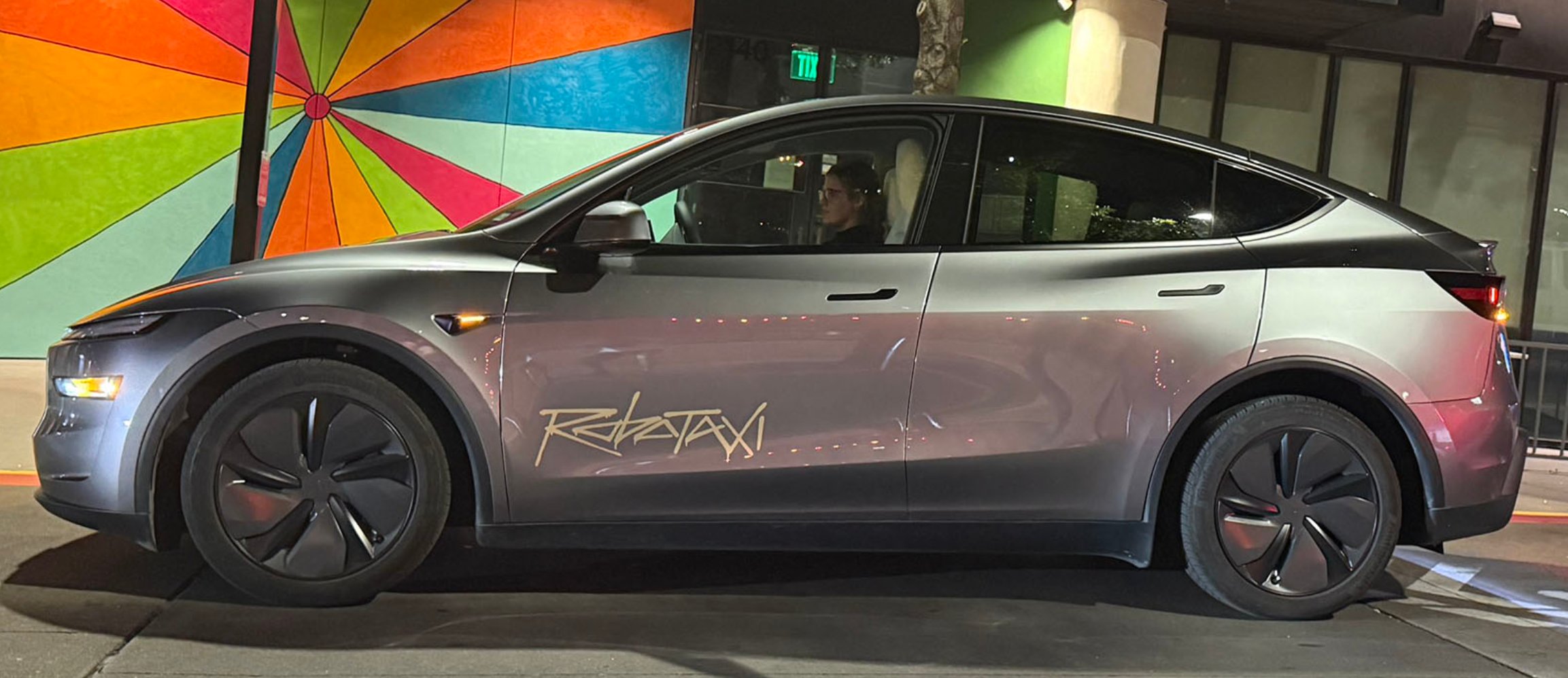 A Tesla Model Y in matte gunmetal with a gold 'Robotaxi' script decal, parked at night in front of a rainbow pinwheel mural.